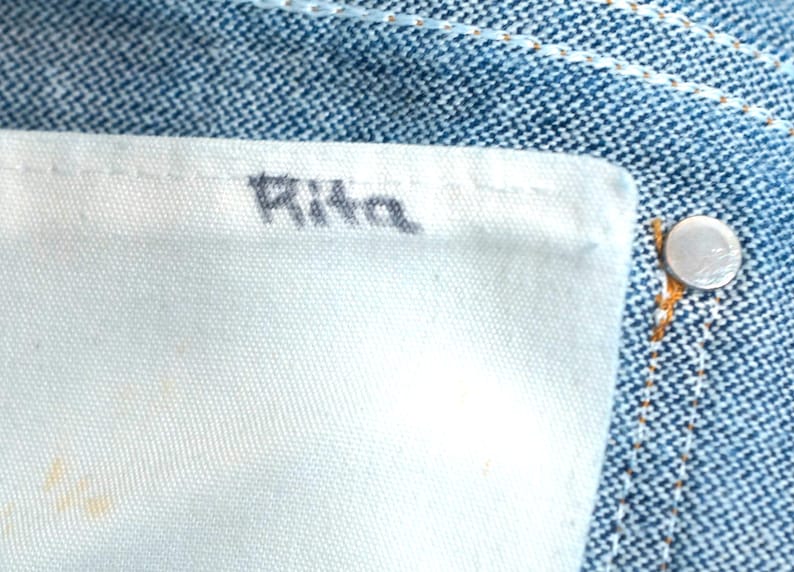 May include: Close-up of denim jeans with a white fabric label. The word "Rita" is printed in black on the label. A silver button is visible on the jeans.