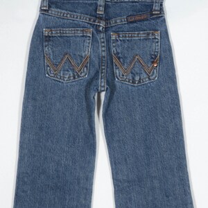 May include: Blue denim jeans with the Wrangler logo on the back pockets. The jeans are hanging on a metal hanger. The stitching is a contrasting brown color.