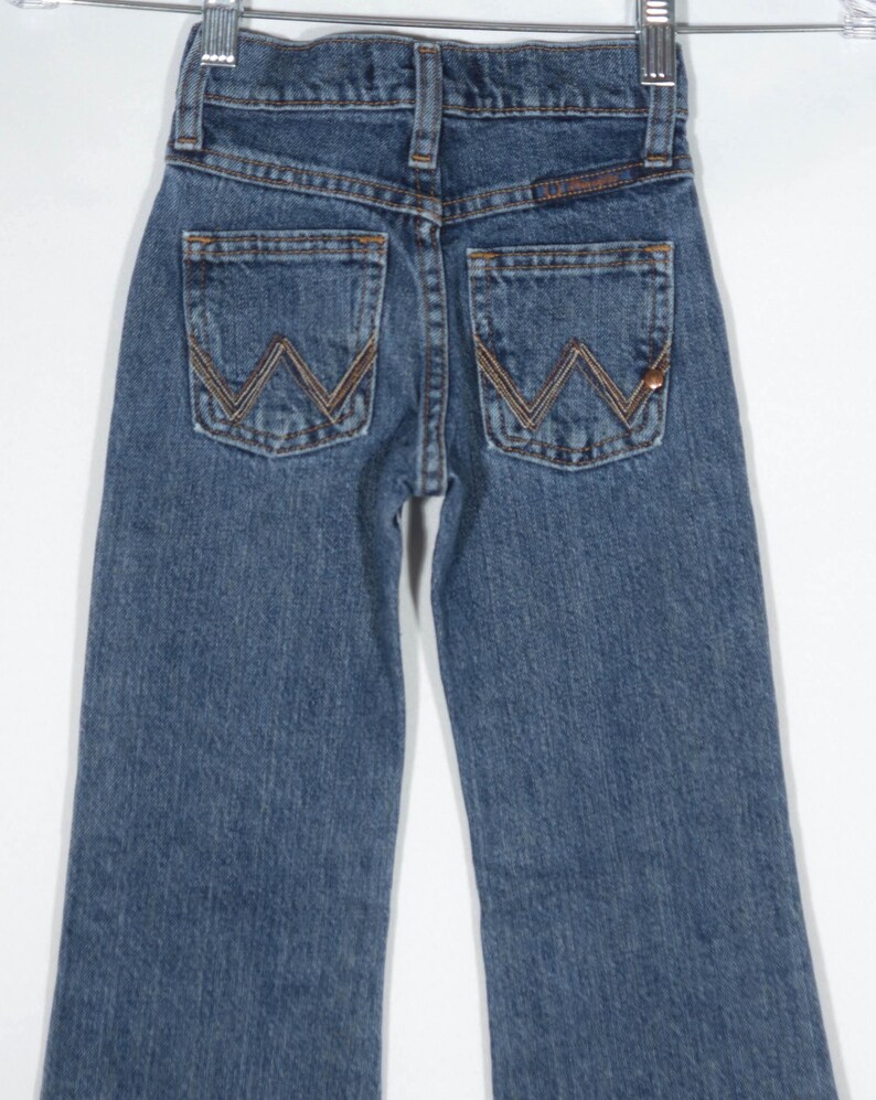 May include: Blue denim jeans with the Wrangler logo on the back pockets. The jeans are hanging on a metal hanger. The stitching is a contrasting brown color.