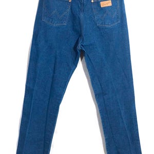 May include: Back view of blue denim jeans hanging on a metal hanger. The jeans have two back pockets with a stitched design, a brown leather patch, and a Wrangler label. The jeans are a medium wash.