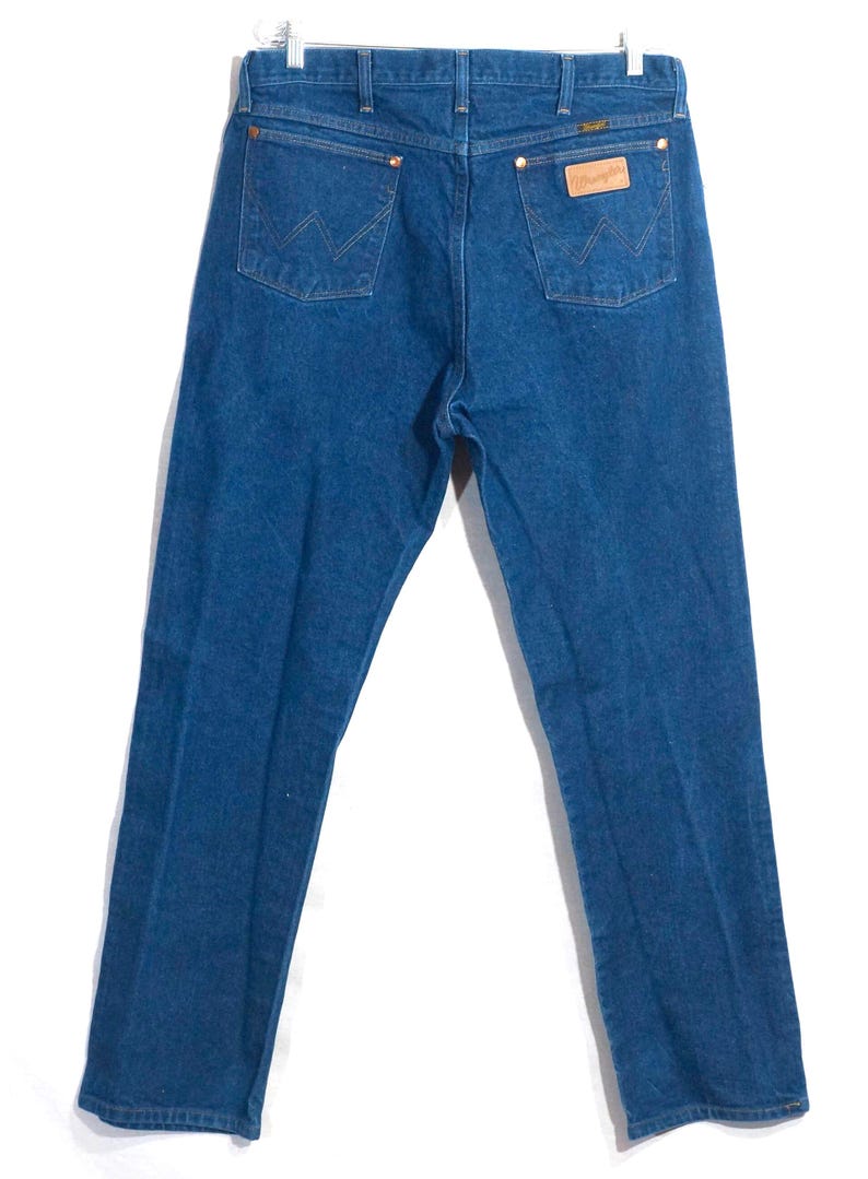 May include: Back view of blue denim jeans hanging on a metal hanger. The jeans have two back pockets with a stitched design, a brown leather patch, and a Wrangler label. The jeans are a medium wash.