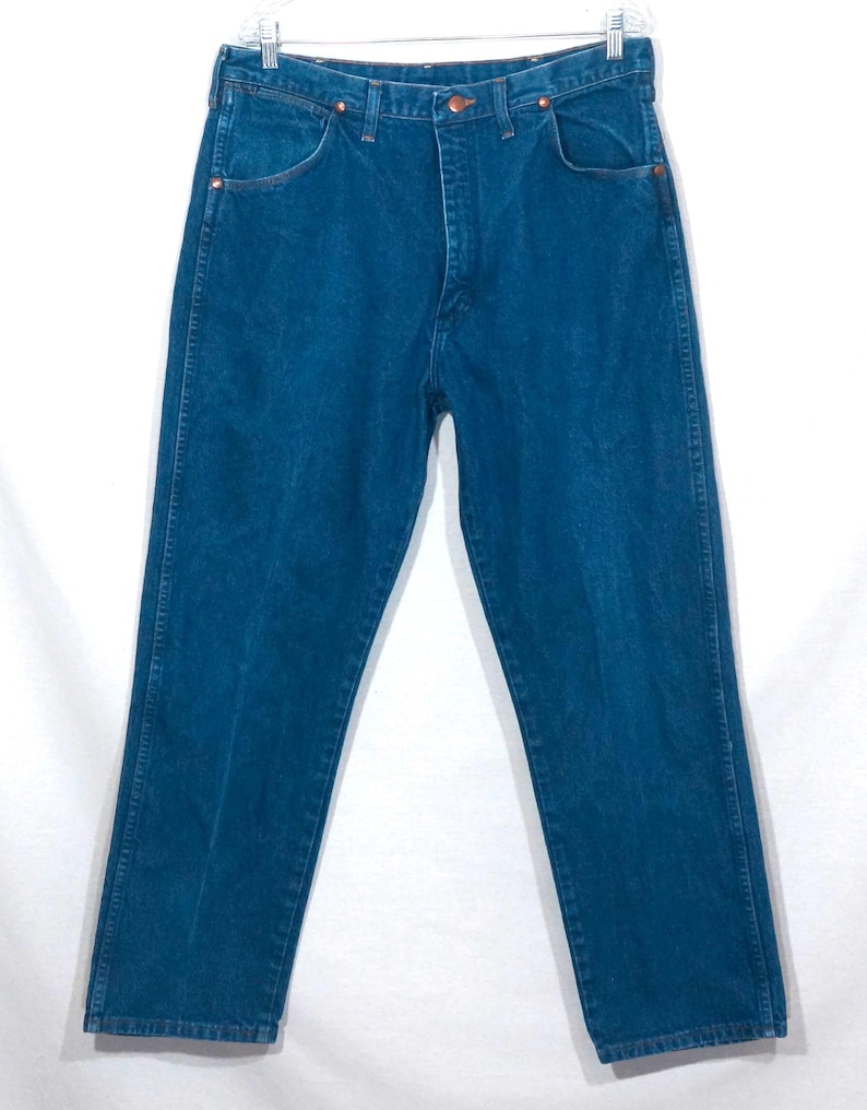 May include: A pair of blue denim jeans, hanging on a white background. The jeans have a classic five-pocket design, with copper rivets and a button closure. The denim is a medium wash, with subtle fading.