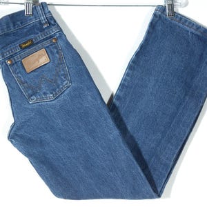 Puede incluir: Vaqueros azules de mezclilla con un corte recto cl&aacute;sico. Los vaqueros tienen un parche de cuero marr&oacute;n en el bolsillo trasero con el logo de Wrangler. Los vaqueros est&aacute;n colgados de una percha de metal.
