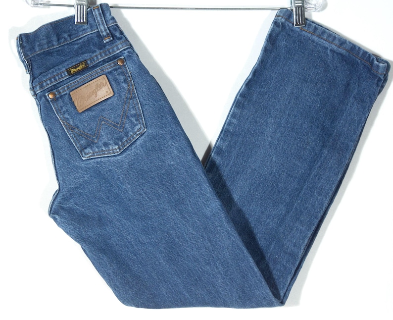 Puede incluir: Vaqueros azules de mezclilla con un corte recto cl&aacute;sico. Los vaqueros tienen un parche de cuero marr&oacute;n en el bolsillo trasero con el logo de Wrangler. Los vaqueros est&aacute;n colgados de una percha de metal.