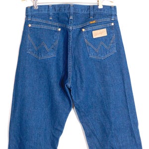 May include: Back view of blue denim jeans with the Wrangler logo on the back pocket. The jeans have a classic five-pocket design with copper-colored rivets and stitching. The jeans are hanging on a hanger.