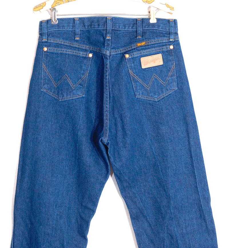 May include: Back view of blue denim jeans with the Wrangler logo on the back pocket. The jeans have a classic five-pocket design with copper-colored rivets and stitching. The jeans are hanging on a hanger.