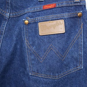 May include: Close-up of the back pocket of blue denim jeans. The pocket features a brown leather patch with the brand name "Wrangler" and a red tag with the brand name. The jeans have copper-coloured rivets and stitching.