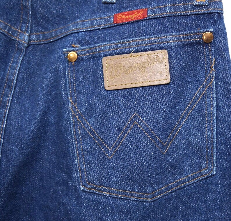 May include: Close-up of the back pocket of blue denim jeans. The pocket features a brown leather patch with the brand name "Wrangler" and a red tag with the brand name. The jeans have copper-coloured rivets and stitching.