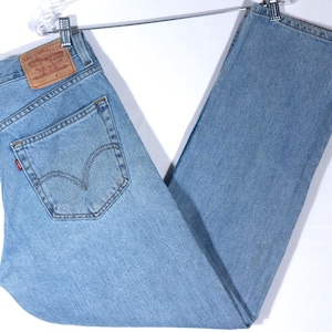 May include: Light blue denim jeans with a classic five-pocket design. The jeans feature a brown leather patch with the brand logo and a small red tag on the back pocket. The jeans are hanging on a metal hanger.