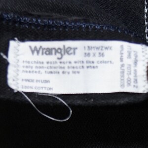 May include: Close-up of a black Wrangler jeans label. The label is white with black text, including the brand name, size 36 x 36, and care instructions. Made in the USA from 100% cotton.