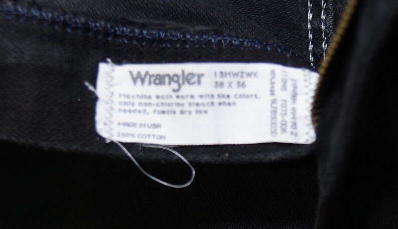 May include: Close-up of a black Wrangler jeans label. The label is white with black text, including the brand name, size 36 x 36, and care instructions. Made in the USA from 100% cotton.