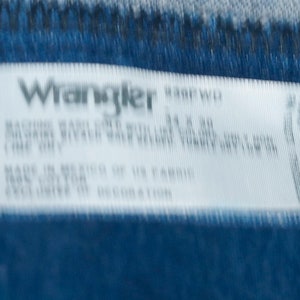 May include: A white label with the Wrangler logo and product information. The label is on a blue denim fabric. The label includes washing instructions and fabric content. The size is 86 x 76 cm.