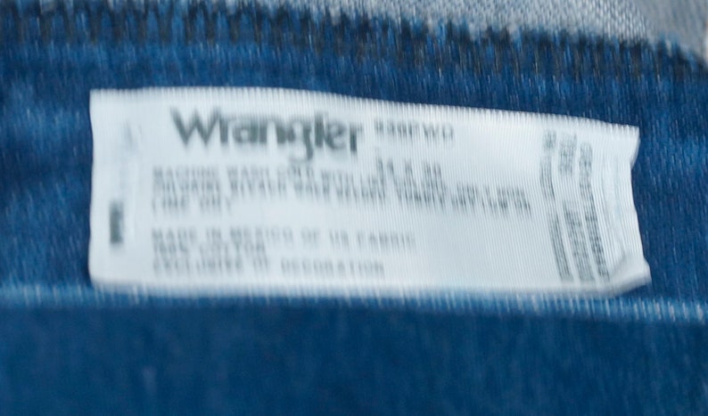 May include: A white label with the Wrangler logo and product information. The label is on a blue denim fabric. The label includes washing instructions and fabric content. The size is 86 x 76 cm.