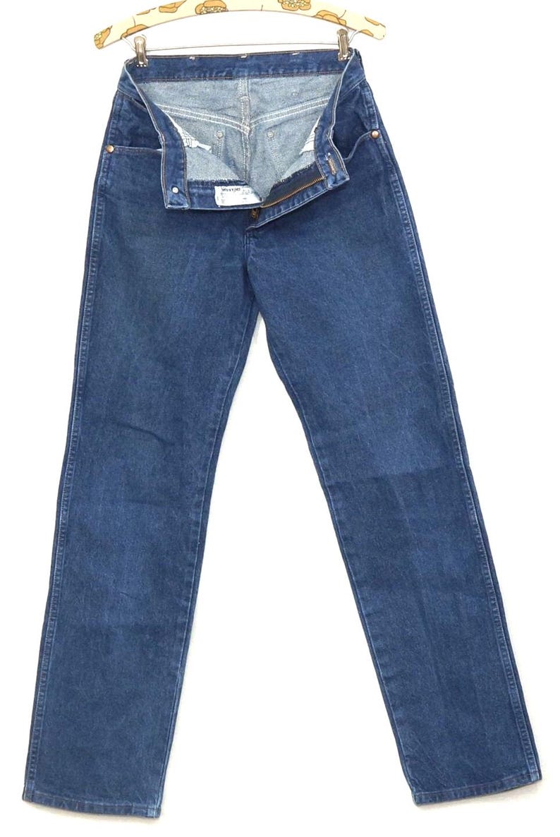 May include: A pair of blue denim jeans, displayed open on a hanger. The jeans have a classic five-pocket design, a button and zipper closure, and a straight leg cut. The denim is a medium wash, and the jeans appear to be vintage.
