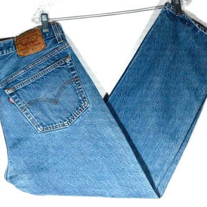May include: Blue denim jeans featuring a tan leather patch on the back waistband with the text Levi Strauss & Co. The jeans have a classic five-pocket design, a red tab on the back pocket, and a medium wash finish, hanging on a metal hanger.