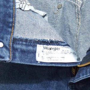 May include: Close-up of blue denim jeans with a visible label. The label reads "Wrangler" and includes washing instructions and size information. The jeans have a zipper and metal button details.