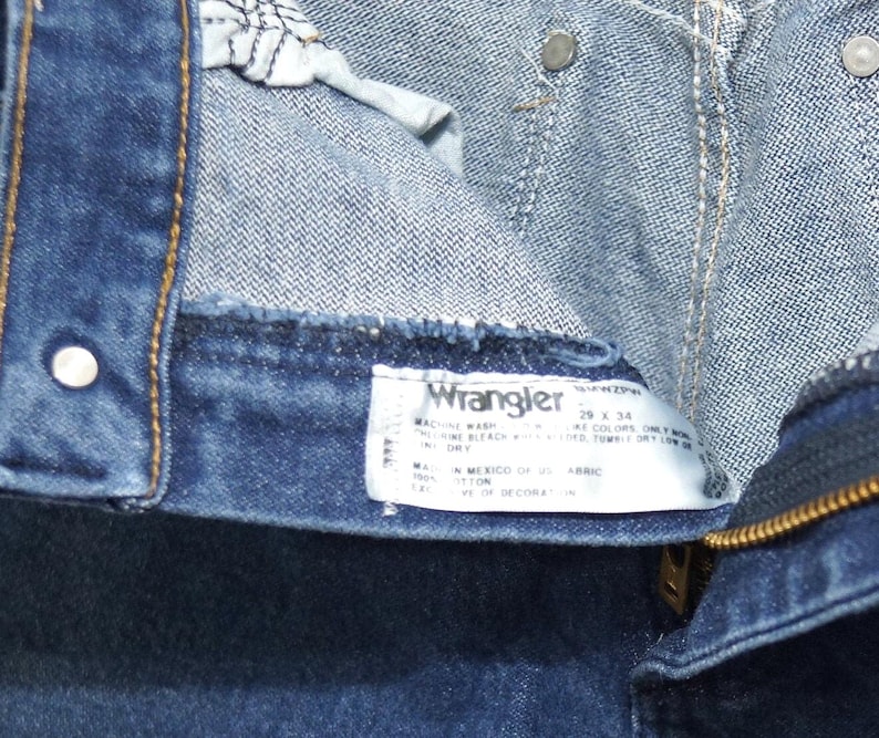 May include: Close-up of blue denim jeans with a visible label. The label reads "Wrangler" and includes washing instructions and size information. The jeans have a zipper and metal button details.