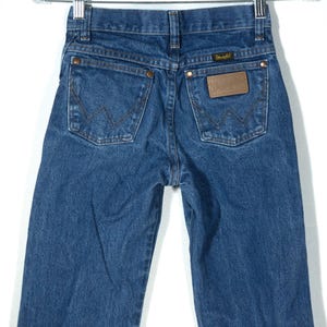 Puede incluir: Vaqueros Wrangler de mezclilla azul con un dise&ntilde;o cl&aacute;sico de cinco bolsillos. Los vaqueros presentan un parche de cuero marr&oacute;n con el logotipo de Wrangler y remaches de cobre. Los bolsillos traseros tienen las costuras distintivas de Wrangler.