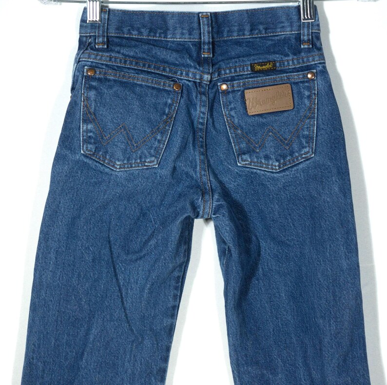 Puede incluir: Vaqueros Wrangler de mezclilla azul con un dise&ntilde;o cl&aacute;sico de cinco bolsillos. Los vaqueros presentan un parche de cuero marr&oacute;n con el logotipo de Wrangler y remaches de cobre. Los bolsillos traseros tienen las costuras distintivas de Wrangler.