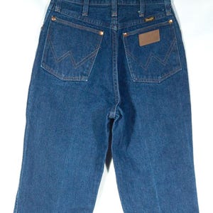 May include: Dark blue denim jeans with the Wrangler logo on the back pocket. The jeans have a classic five-pocket design with copper rivets and a brown leather patch. The jeans are hanging on a metal hanger.