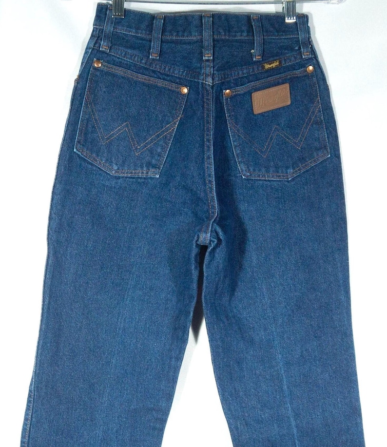 May include: Dark blue denim jeans with the Wrangler logo on the back pocket. The jeans have a classic five-pocket design with copper rivets and a brown leather patch. The jeans are hanging on a metal hanger.