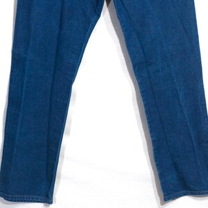 May include: Close-up of blue denim jeans. The jeans are a medium wash, with a classic five-pocket design. The image shows the legs of the jeans, with the fabric appearing slightly faded in areas. The jeans are likely vintage or pre-owned.