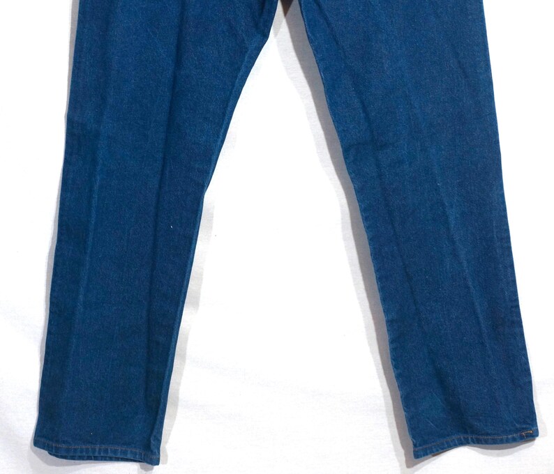 May include: Close-up of blue denim jeans. The jeans are a medium wash, with a classic five-pocket design. The image shows the legs of the jeans, with the fabric appearing slightly faded in areas. The jeans are likely vintage or pre-owned.