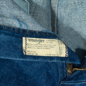 May include: Close-up of blue denim Wrangler jeans. The tag reads "Wrangler 8 x 32" and "Made in USA 100% Cotton." The zipper is visible, along with the top of the jeans and the button closure.