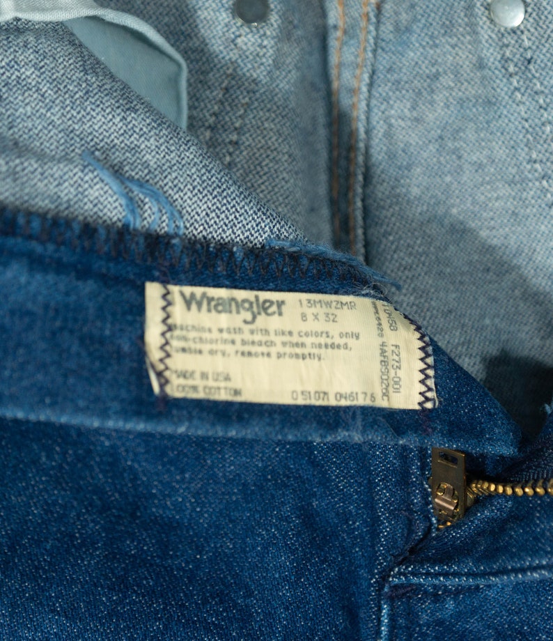 May include: Close-up of blue denim Wrangler jeans. The tag reads "Wrangler 8 x 32" and "Made in USA 100% Cotton." The zipper is visible, along with the top of the jeans and the button closure.