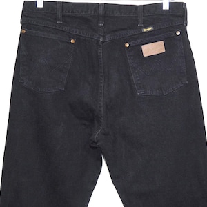May include: Black denim jeans with classic five-pocket styling. Features include brass rivets, a leather patch, and the Wrangler logo. The back pockets have the signature W stitching.