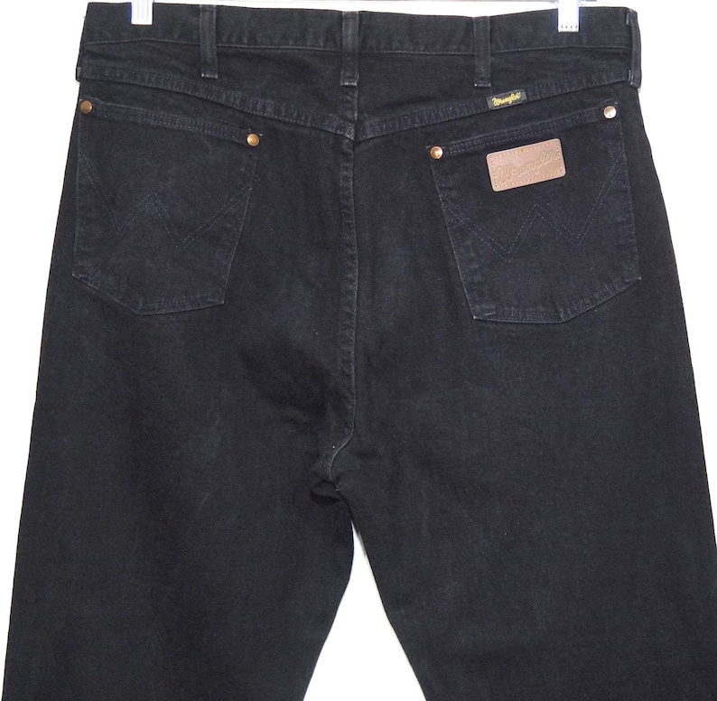 May include: Black denim jeans with classic five-pocket styling. Features include brass rivets, a leather patch, and the Wrangler logo. The back pockets have the signature W stitching.