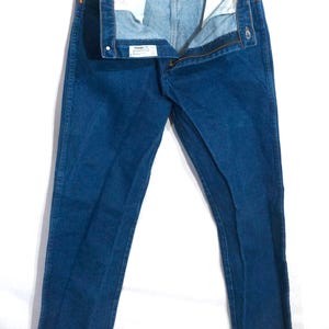 May include: A pair of blue denim jeans, hanging open, with a visible zipper and button closure. The jeans are a medium wash, with a classic five-pocket design. The interior of the waistband is visible, showing the brand label.