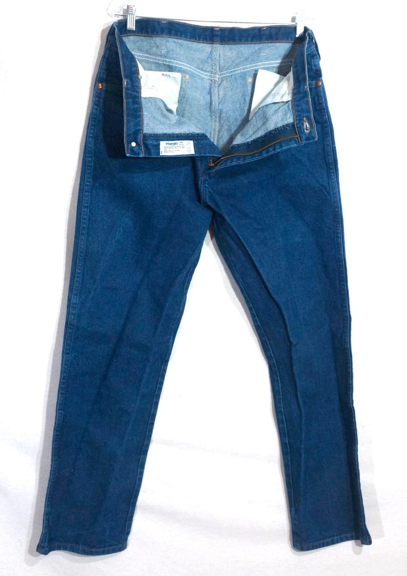 May include: A pair of blue denim jeans, hanging open, with a visible zipper and button closure. The jeans are a medium wash, with a classic five-pocket design. The interior of the waistband is visible, showing the brand label.