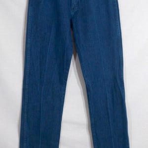 May include: A pair of classic blue denim jeans hanging on a clear hanger. The jeans have a traditional five-pocket design with copper-coloured rivets and a button closure. The denim is a medium wash, showing subtle fading.