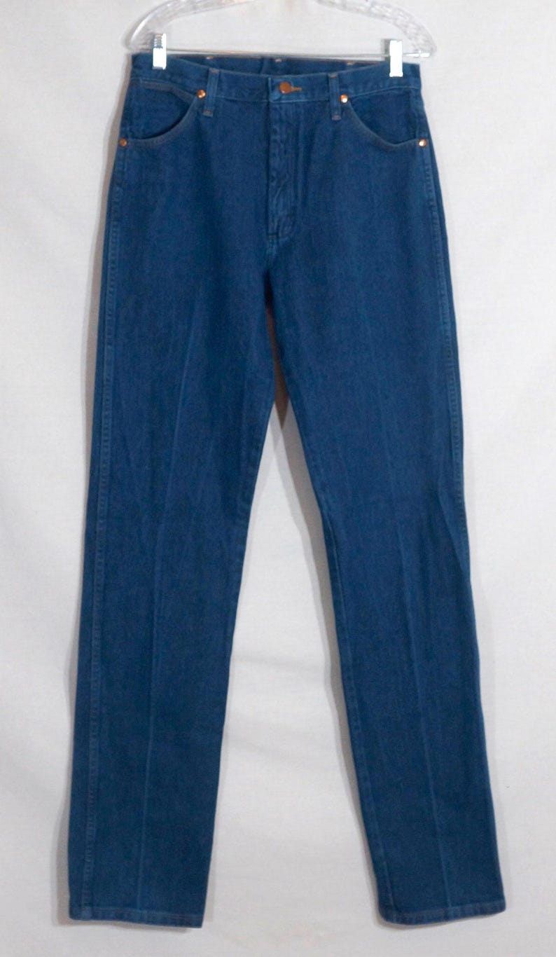 May include: A pair of classic blue denim jeans hanging on a clear hanger. The jeans have a traditional five-pocket design with copper-coloured rivets and a button closure. The denim is a medium wash, showing subtle fading.