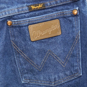 May include: Close-up of the back pocket of blue denim jeans. The pocket features a brown leather patch with the embossed word "Wrangler". The pocket is outlined with tan stitching, and the jeans have copper rivets.