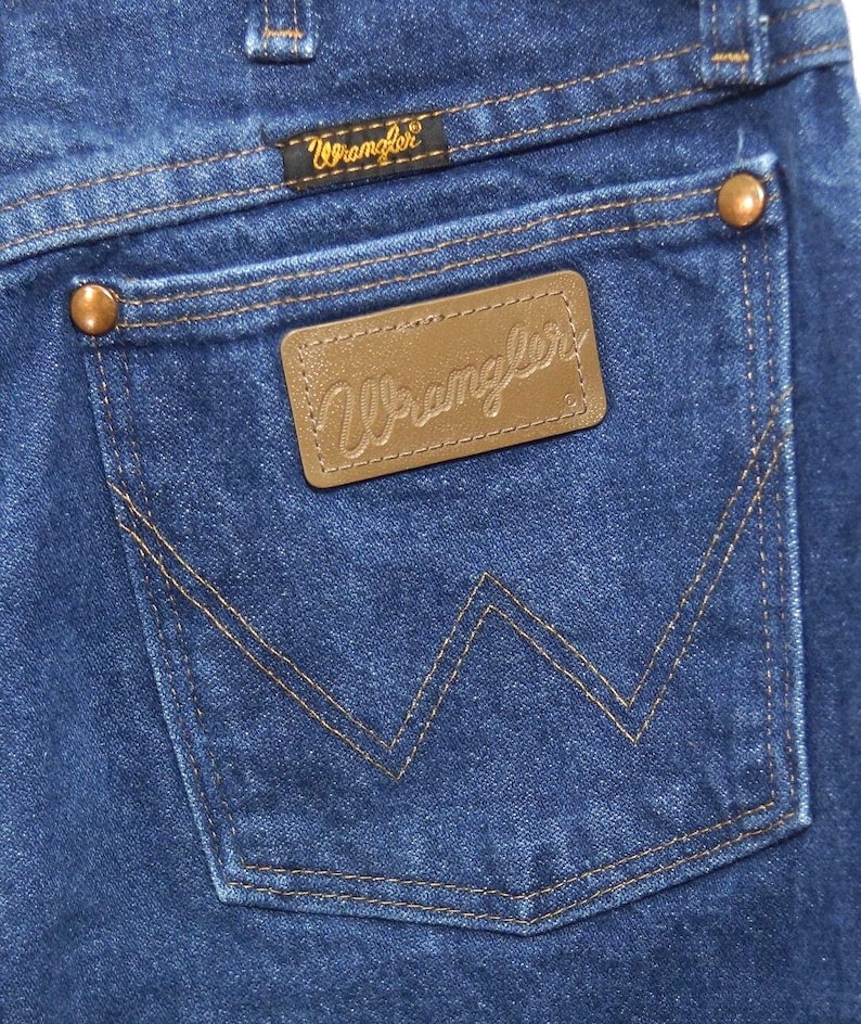 May include: Close-up of the back pocket of blue denim jeans. The pocket features a brown leather patch with the embossed word "Wrangler". The pocket is outlined with tan stitching, and the jeans have copper rivets.