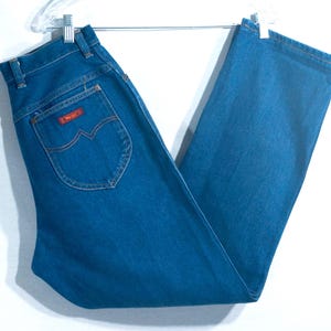 May include: Blue denim jeans with a classic straight leg cut. The jeans feature a red "Wrangler" patch on the back pocket and are hanging on a metal hanger. The jeans are a medium wash and appear to be vintage.