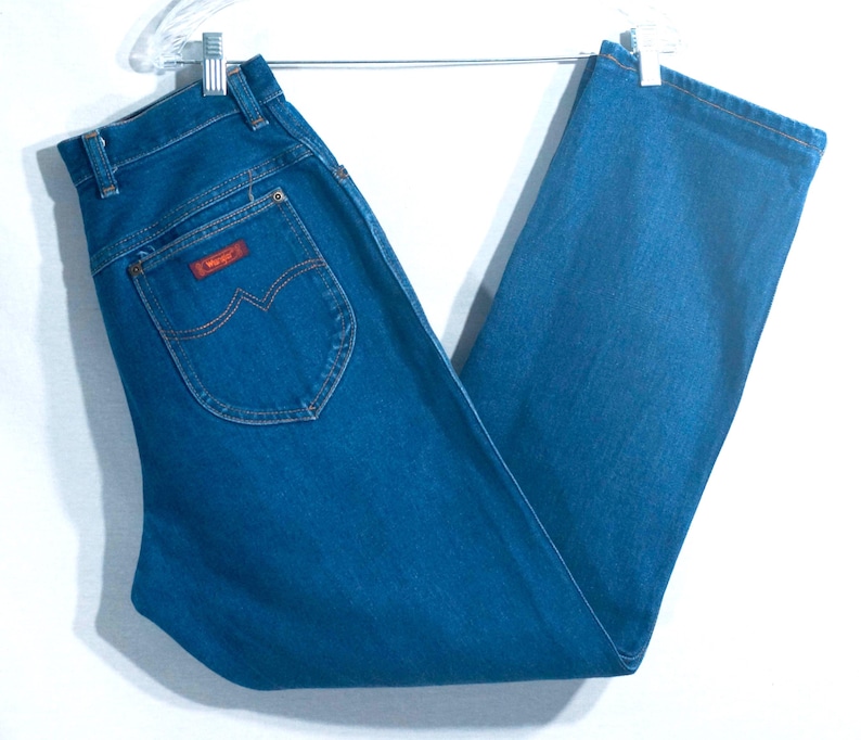 May include: Blue denim jeans with a classic straight leg cut. The jeans feature a red "Wrangler" patch on the back pocket and are hanging on a metal hanger. The jeans are a medium wash and appear to be vintage.