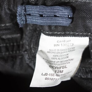 May include: Close-up of black denim pants with an adjustable elastic waistband. A white tag displays the text "CARE007 RN 130273 MADE IN BANGLADESH 100% COTTON 12M". The pants are made of 100% cotton.