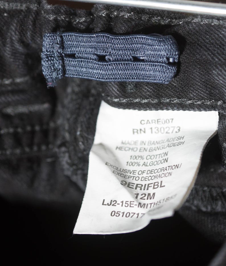 May include: Close-up of black denim pants with an adjustable elastic waistband. A white tag displays the text "CARE007 RN 130273 MADE IN BANGLADESH 100% COTTON 12M". The pants are made of 100% cotton.