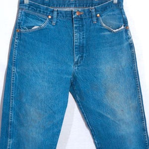 Puede incluir: Vaqueros azules de mezclilla con un dise&ntilde;o cl&aacute;sico de cinco bolsillos. Los vaqueros tienen cierre de bot&oacute;n y cremallera, remaches de cobre y un lavado descolorido. Los vaqueros est&aacute;n colgados de una percha de metal.