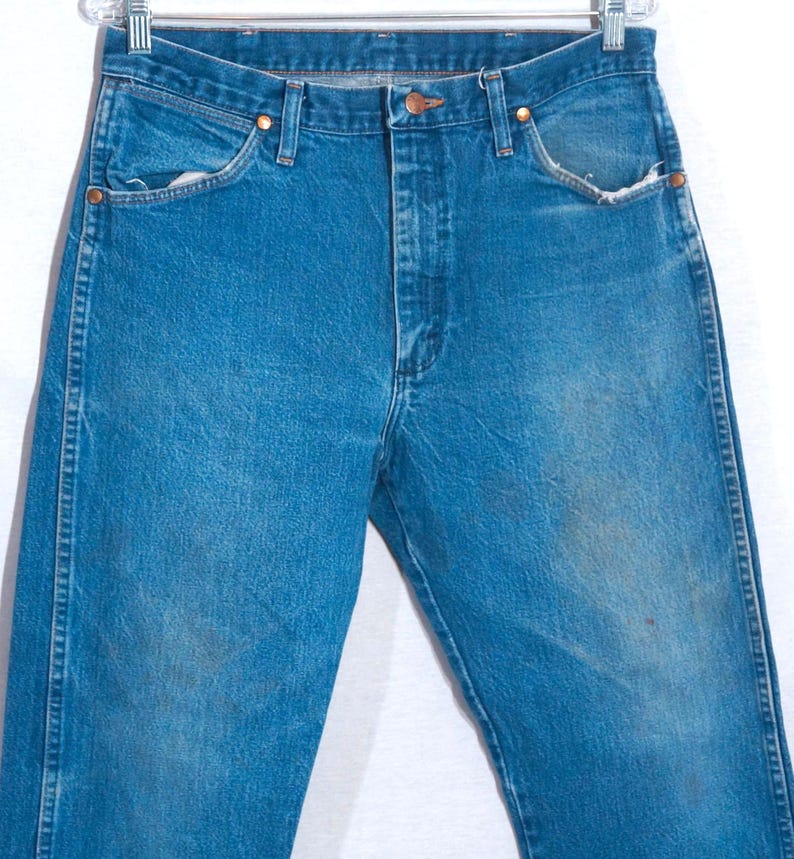 Puede incluir: Vaqueros azules de mezclilla con un dise&ntilde;o cl&aacute;sico de cinco bolsillos. Los vaqueros tienen cierre de bot&oacute;n y cremallera, remaches de cobre y un lavado descolorido. Los vaqueros est&aacute;n colgados de una percha de metal.