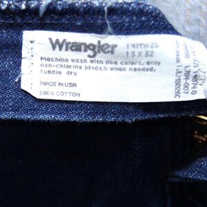 May include: Close-up of dark blue denim fabric with a white label. The label reads "Wrangler" and includes washing instructions and size details: 13x32. The tag also states "MADE IN USA" and "100% COTTON".