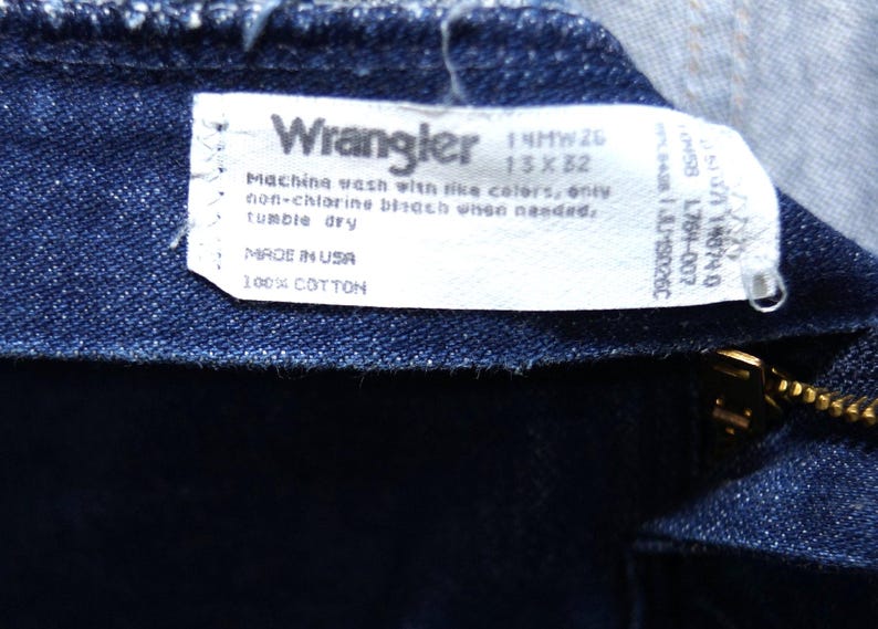 May include: Close-up of dark blue denim fabric with a white label. The label reads "Wrangler" and includes washing instructions and size details: 13x32. The tag also states "MADE IN USA" and "100% COTTON".
