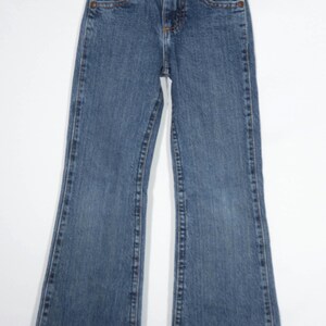 May include: Blue denim jeans with a classic five-pocket design and a flared leg. The jeans have a button and zipper closure, along with copper-colored rivets and stitching details. The jeans are hanging on a white hanger.