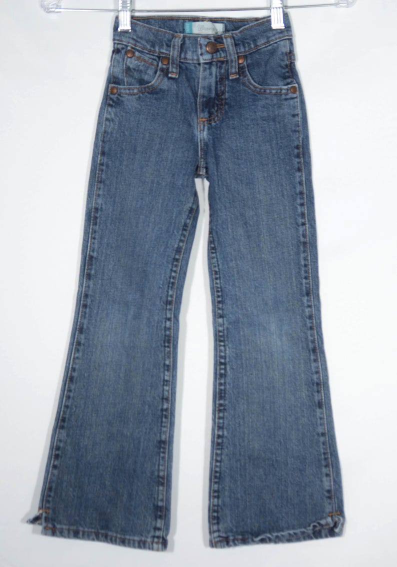 May include: Blue denim jeans with a classic five-pocket design and a flared leg. The jeans have a button and zipper closure, along with copper-colored rivets and stitching details. The jeans are hanging on a white hanger.
