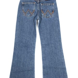 May include: Blue denim jeans with a classic five-pocket design. The back pockets feature a stitched "W" design in a contrasting brown thread. The jeans have a flared leg and are hanging on a hanger.