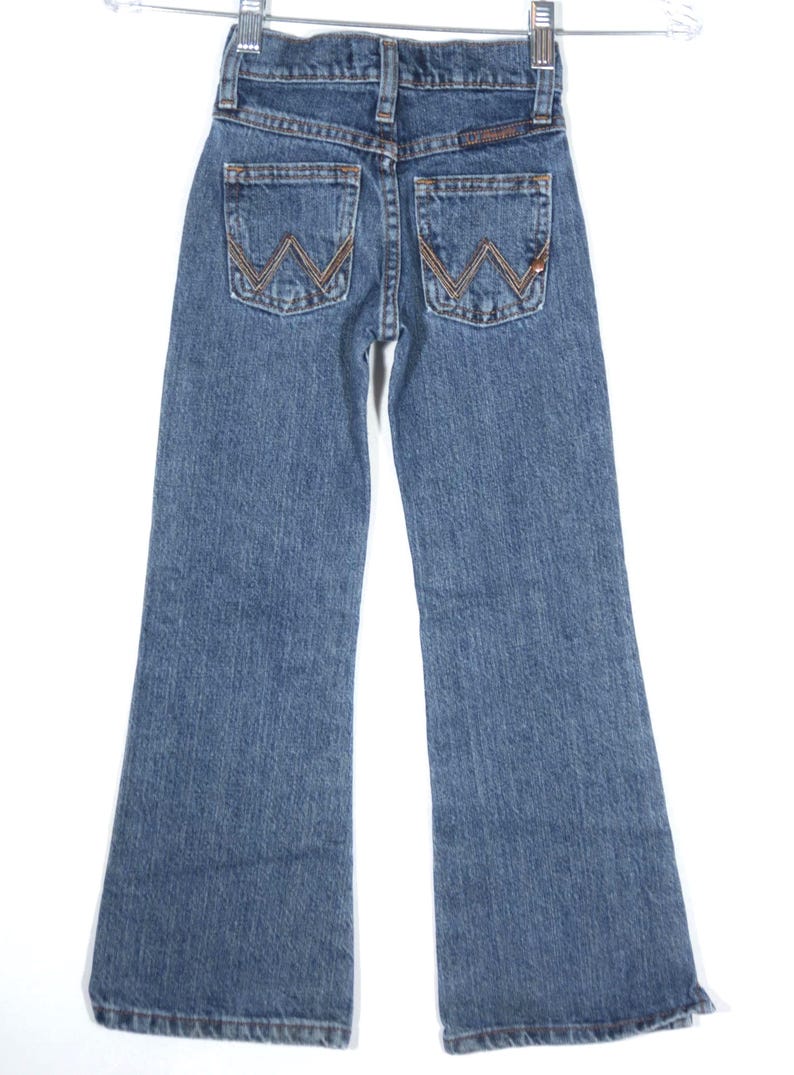 May include: Blue denim jeans with a classic five-pocket design. The back pockets feature a stitched "W" design in a contrasting brown thread. The jeans have a flared leg and are hanging on a hanger.