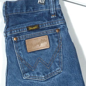 Puede incluir: Primer plano de unos vaqueros de mezclilla azules con un parche de cuero marr&oacute;n en el bolsillo trasero. El parche y la etiqueta peque&ntilde;a llevan el nombre de la marca "Wrangler". Los vaqueros cuelgan de una percha met&aacute;lica.